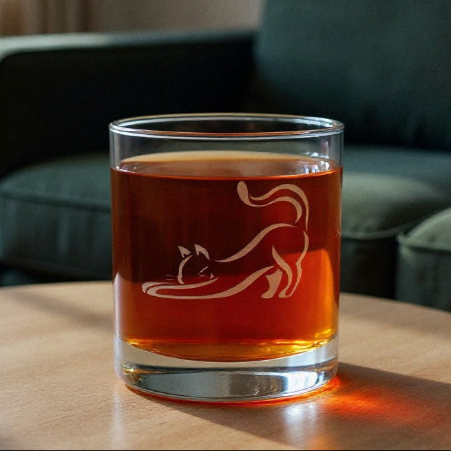 A clear rocks glass with an etched cat silhouette, filled with amber-colored liquid, sitting on a round wooden table in a living room with a green sofa in the background.