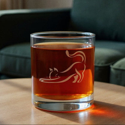 A clear rocks glass with an etched cat silhouette, filled with amber-colored liquid, sitting on a round wooden table in a living room with a green sofa in the background.