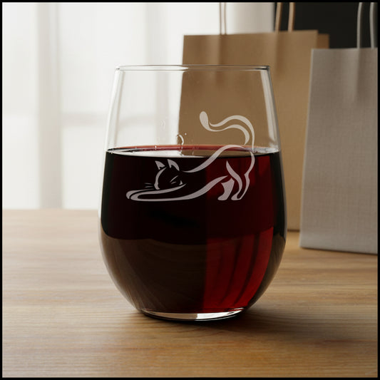 A clear stemless wine glass with an etched black cat silhouette, displayed on a wooden surface gift bags in thebackground.