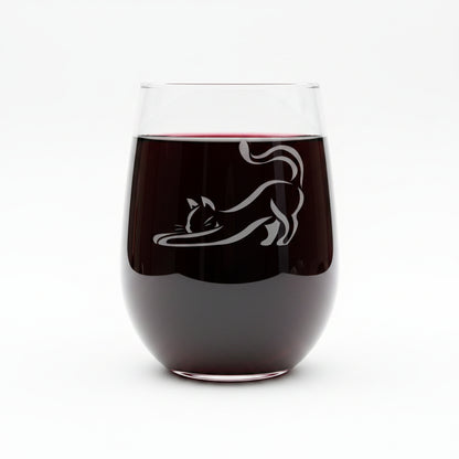 A clear stemless wine glass with an etched black cat silhouette, displayed on a wooden surface against a solid whitebackground.