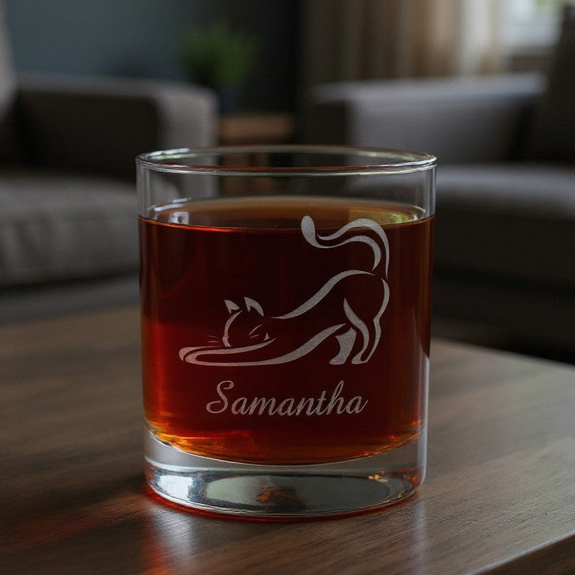A clear rocks glass with an etched cat silhouette and the name 'Samantha', filled with amber liquid, sitting on a wooden table in a softly blurred living room setting.