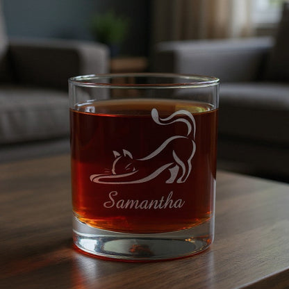 A clear rocks glass with an etched cat silhouette and the name 'Samantha', filled with amber liquid, sitting on a wooden table in a softly blurred living room setting.