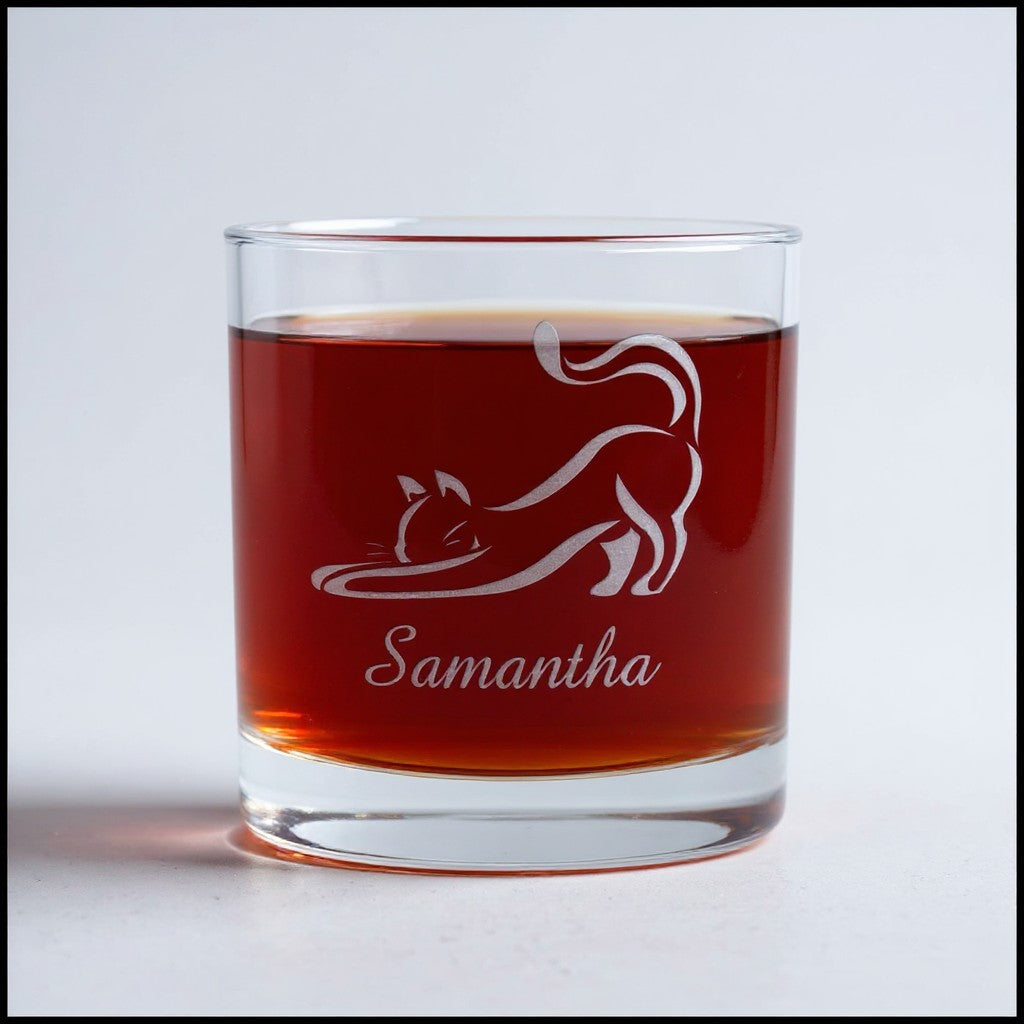 A clear glass rocks tumbler filled with an amber-brown liquid, featuring a white frosted graphic of a stretching cat and the name 'Samantha' printed below. The glass is centered on a light gray background.