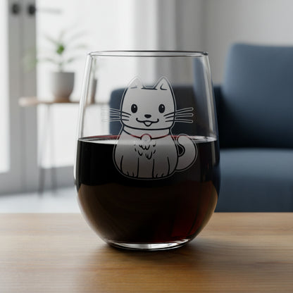 A clear stemless wine glass with aan etched cute cat illustration on the front, set on a wooden surface against a blue couch background.