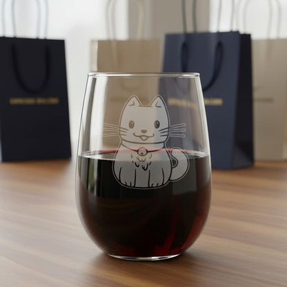 A stemless wine glass filled with red wine sits on a wooden table, featuring an etched cute cat design. In the background, black and beige gift bags are softly blurred.