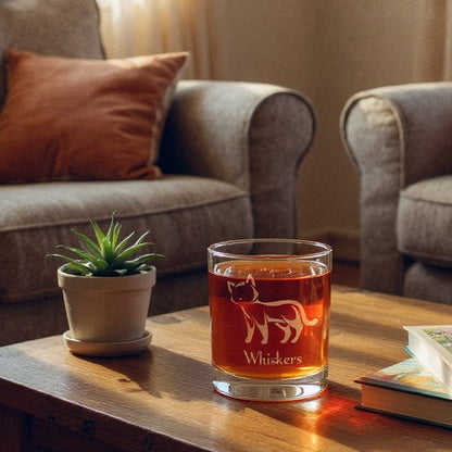 A clear lowball glass with a white cat silhouette and the word 'Whiskers' printed on it, filled with amber liquid, placed on a wooden coffee table in a cozy living room setting with two gray armchairs and orange pillows in the background.