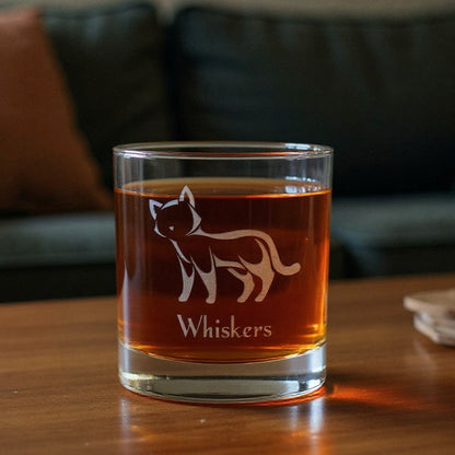 A clear rocks glass with an etched cat silhouette and the name 'Whiskers' sits filled with amber whiskey on a wooden table in a living room setting with a couch in the background.