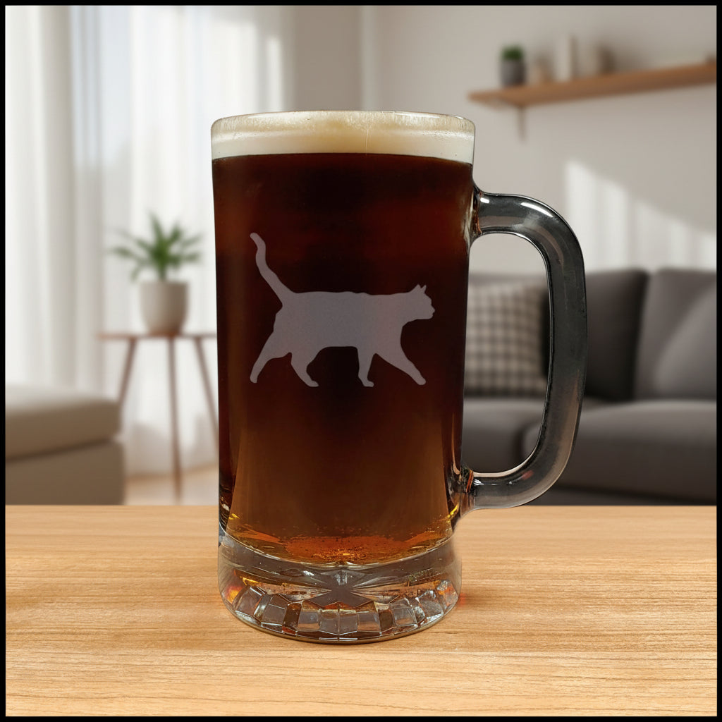 A clear glass beer mug filled with dark beer and topped with white foam, featuring a cat silhouette etched on the front, set on a light wood surface against a living room background.
