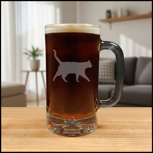 A clear glass beer mug filled with dark beer and topped with white foam, featuring a cat silhouette etched on the front, set on a light wood surface against a living room background.