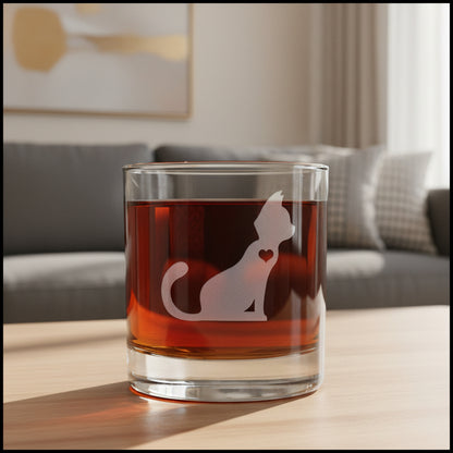 A clear glass rocks tumbler filled with amber-red liquid, featuring a frosted pale pink cat silhouette with a small heart on the side, with a cozy living room in the background.