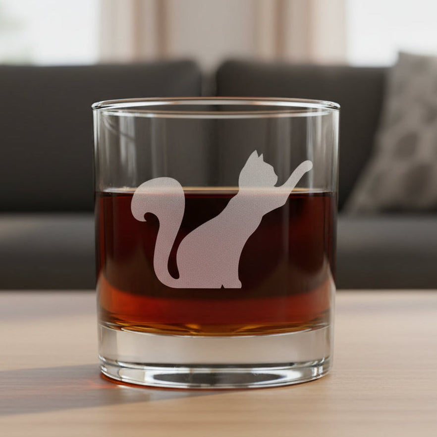A clear rocks glass filled with amber-red liquid, featuring an etched silhouette of a stretching cat on the front, set against a textured gray background.
