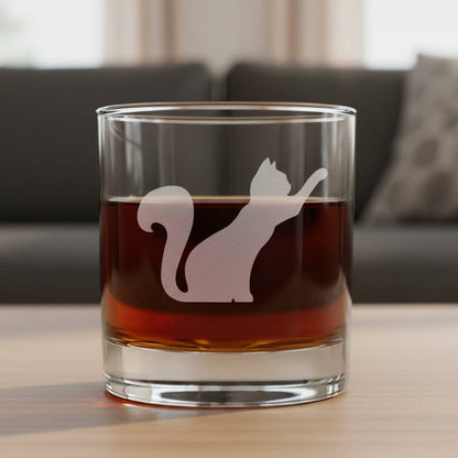 A clear rocks glass filled with amber-red liquid, featuring an etched silhouette of a stretching cat on the front, set against a textured gray background.