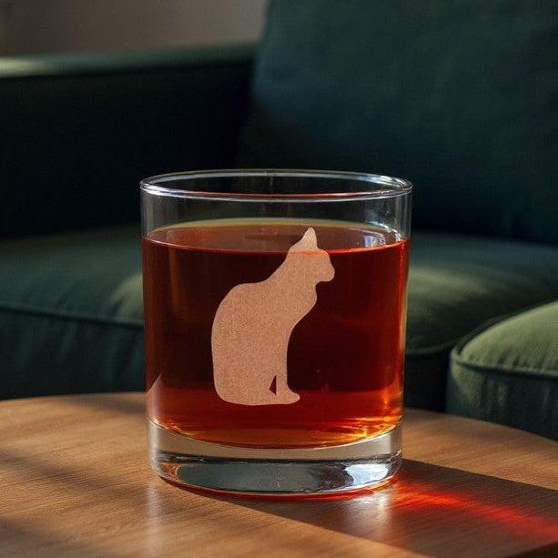 A clear rocks glass with an etched cat silhouette design, filled with an amber drink, set on a round wooden table in a living room with a green sofa in the background.