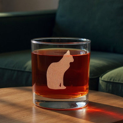 A clear rocks glass with an etched cat silhouette design, filled with an amber drink, set on a round wooden table in a living room with a green sofa in the background.