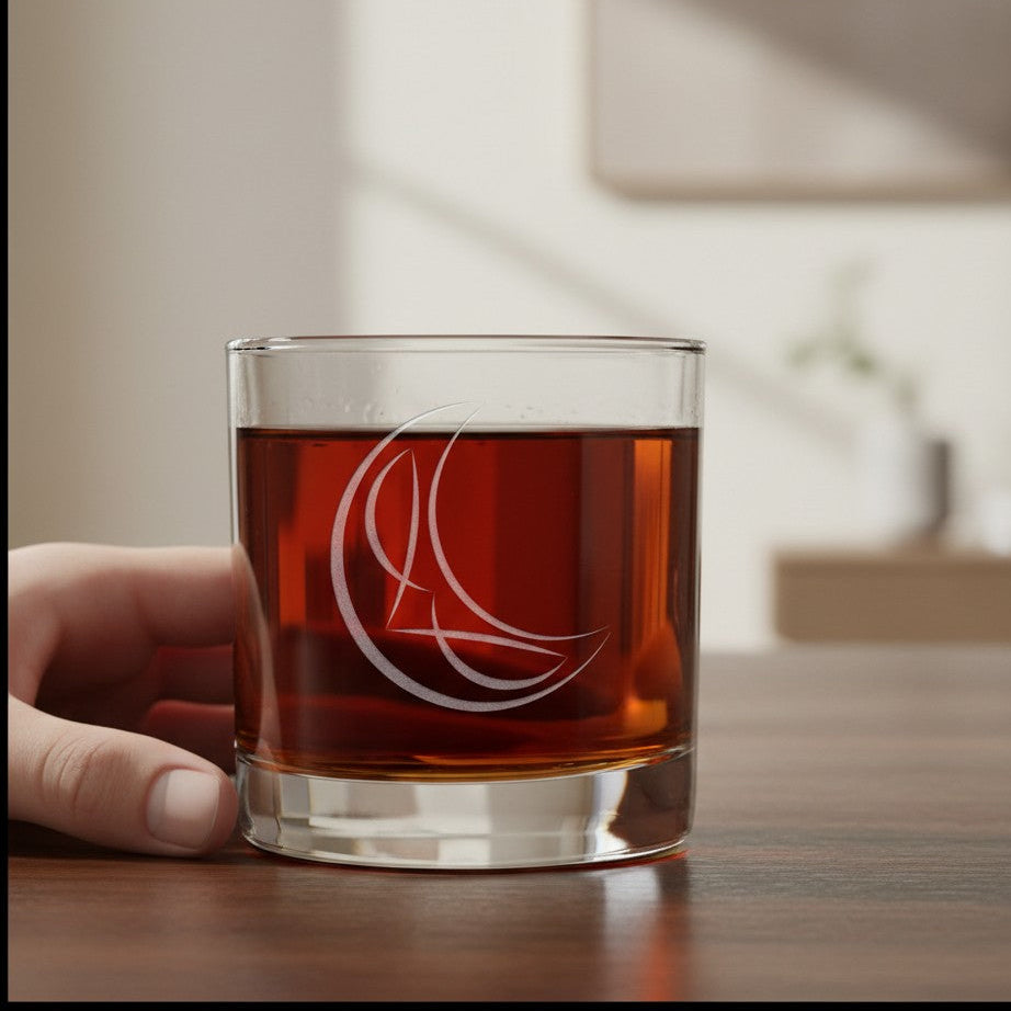 A hand holding a clear rocks glass with an etched creascent moon design, filled with an amber-brown liquid, set on a wooden table in a softly blurred indoor environment.