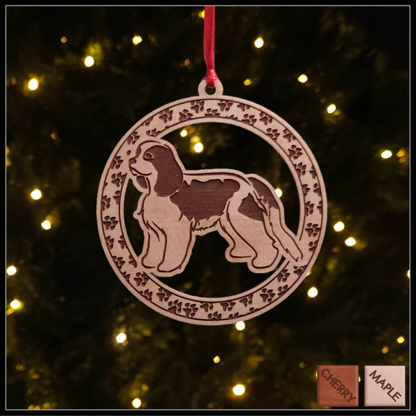 A round maple wood veneer ornament with a border of small paw prints. The center of the ornament is a Cavalier King Charles dog.