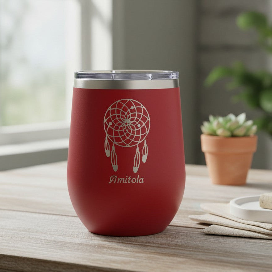 A red insulated stemless wine tumbler with a clear lid sits on a wooden table in a softly lit room. The tumbler has a white dreamcatcher graphic and the name 'Amitola' printed on the front.