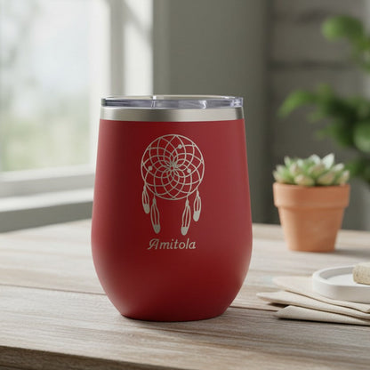 A red insulated stemless wine tumbler with a clear lid sits on a wooden table in a softly lit room. The tumbler has a white dreamcatcher graphic and the name 'Amitola' printed on the front.