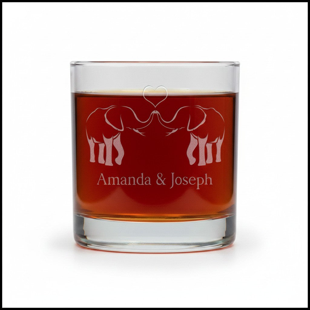 A clear glass rocks tumbler filled with amber liquid, featuring an etched design of two elephants facing each other with a heart above them and the personalized text 'Amanda & Joseph' below, shown on a white background.