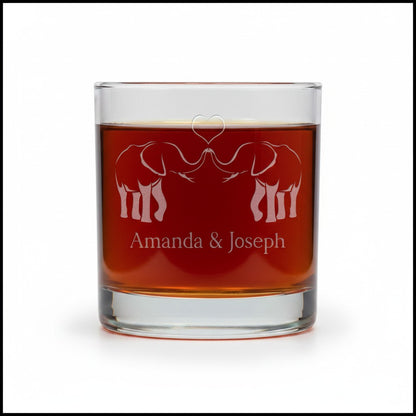 A clear glass rocks tumbler filled with amber liquid, featuring an etched design of two elephants facing each other with a heart above them and the personalized text 'Amanda & Joseph' below, shown on a white background.