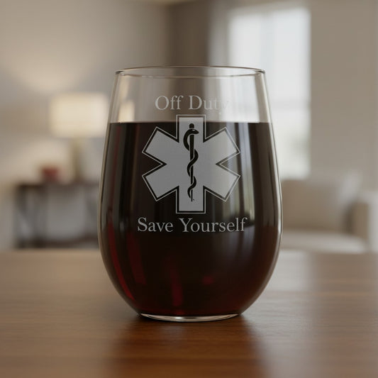 A clear stemless wine glass filled with dark red wine sits on a wooden table in a softly blurred indoor setting. The glass has etched  text reading 'Off Duty' and 'Save Yourself' with a large EMS symbol in the center.