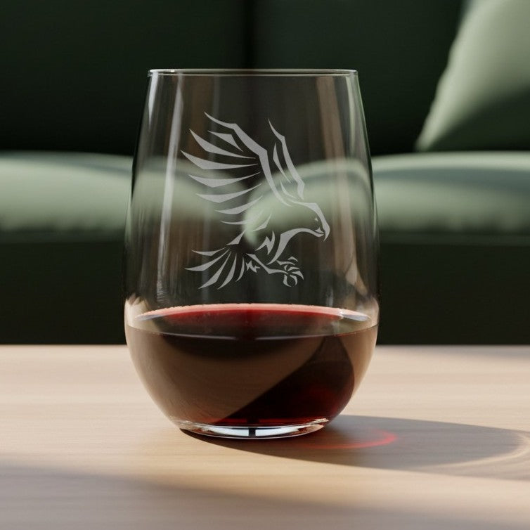 A clear stemless wine glass with an etched eagle design, partially filled with red wine, set on a light wooden table with a blurred dark green sofa in the background.