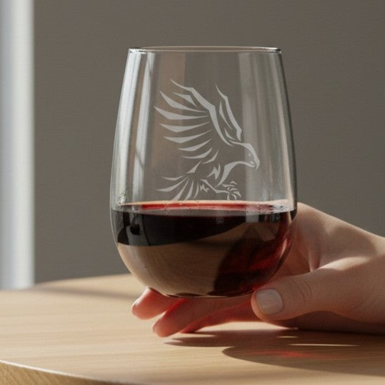 A hand holding a clear stemless wine glass with an etched eagle design, partially filled with red wine, set on a light wooden table against a soft gray background.