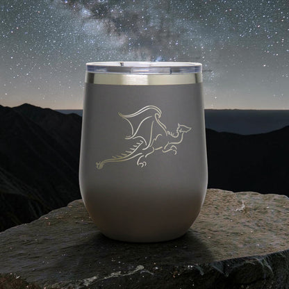 A matte gray insulated stemless tumbler with a clear lid sits on a dark stone surface against a night sky backdrop with mountains and stars. The tumbler is etched with a dragon design.