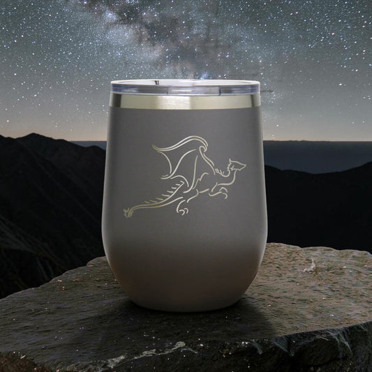 A matte gray insulated stemless tumbler with a clear lid sits on a dark stone surface against a night sky backdrop with mountains and stars. The tumbler is etched with a dragon design.