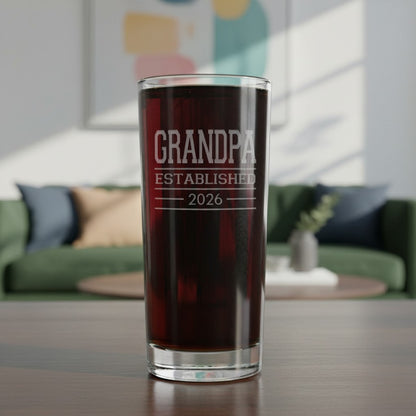 A clear glass tumbler with 'GRANDPA ESTABLISHED 2026' etched on it  with a very dark red liquid, set on a wooden table in a blurred living room background.
