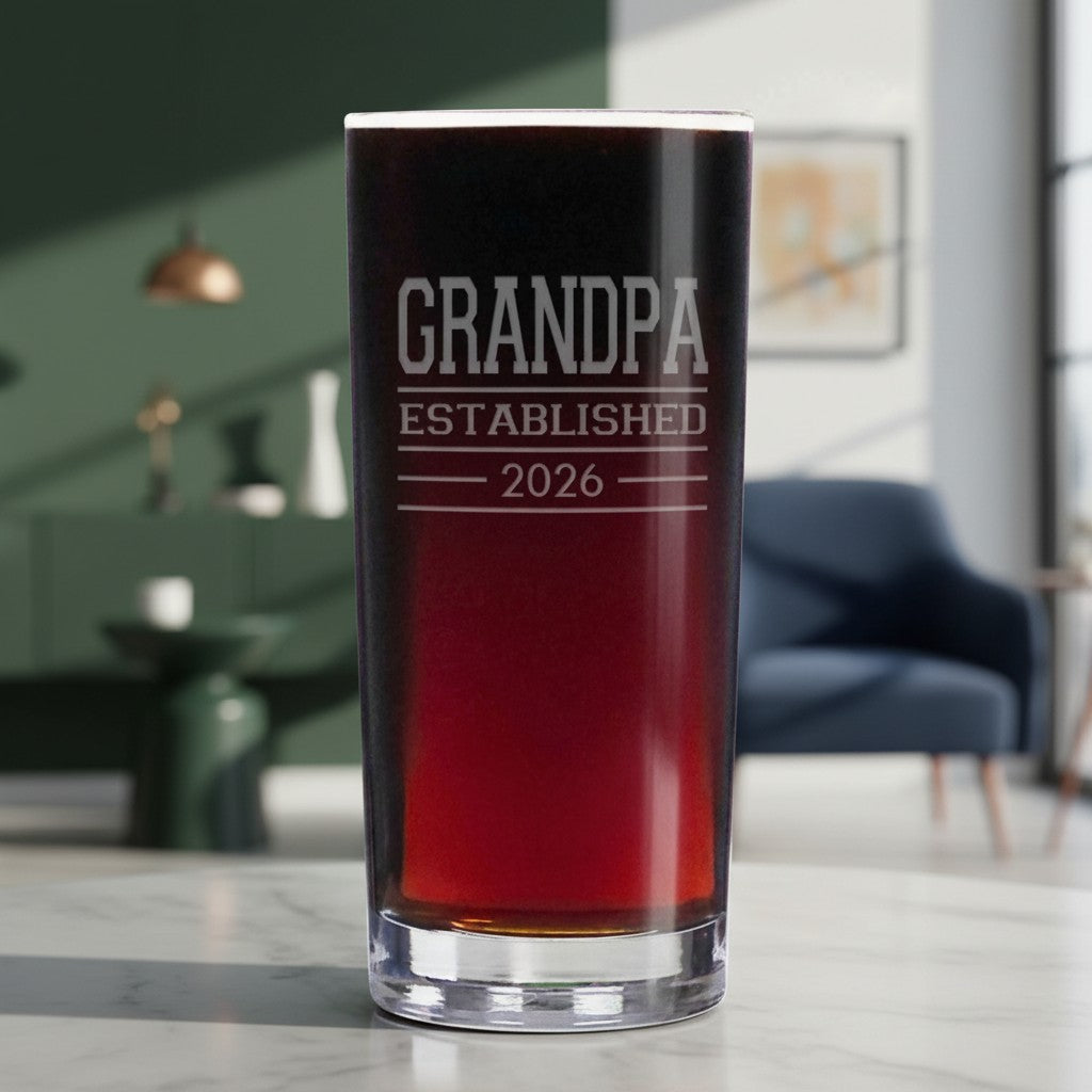 A clear glass cooler glass filled with a dark red drink, standing on a light marble countertop in a blurred living room. The glass is etched with the text 'GRANDPA ESTABLISHED 2026'.