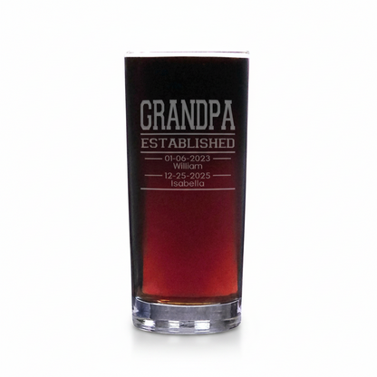 A clear glass shot glass filled with a dark red liquid, featuring custom white etched text that reads 'GRANDPA ESTABLISHED' with dates and names below, set against a plain white background.