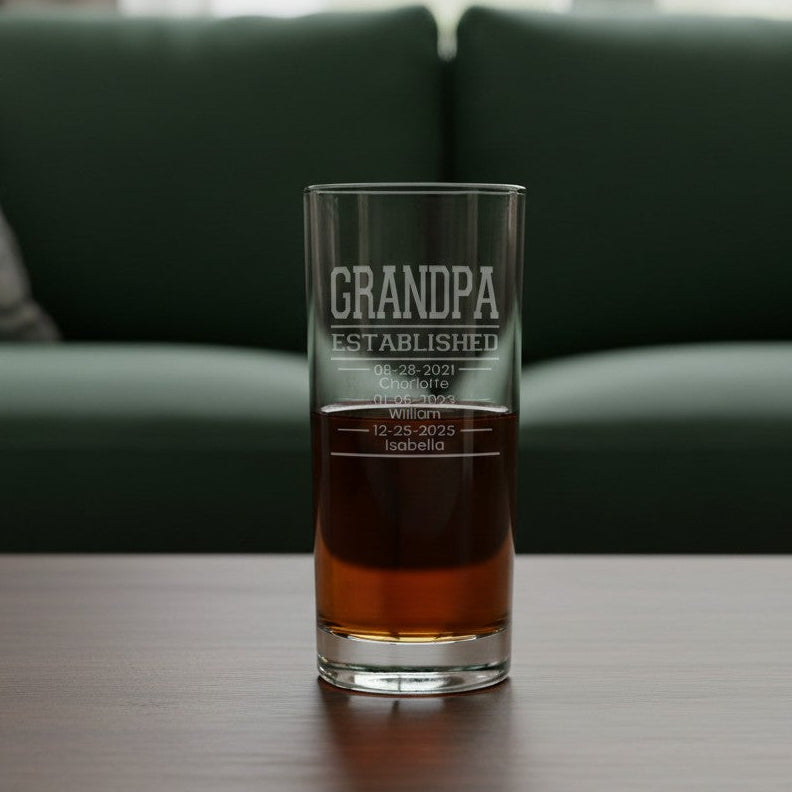 A clear glass cooler glass with 'GRANDPA ESTABLISHED' etched on the front, partially filled with an amber drink, sitting on a wooden table in front of a green sofa.