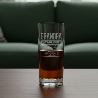 A clear glass cooler glass with 'GRANDPA ESTABLISHED' etched on the front, partially filled with an amber drink, sitting on a wooden table in front of a green sofa.