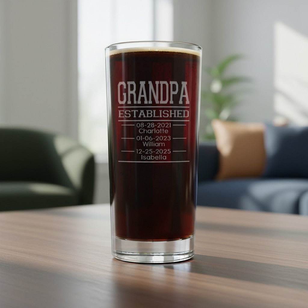 A clear cooler glass filled with dark beer, standing on a wooden table in a softly blurred living room. The glass is etched with the text 'GRANDPA ESTABLISHED' followed by personalized dates and names.