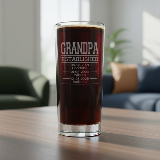 A clear cooler glass filled with dark beer, standing on a wooden table in a softly blurred living room. The glass is etched with the text 'GRANDPA ESTABLISHED' followed by personalized dates and names.