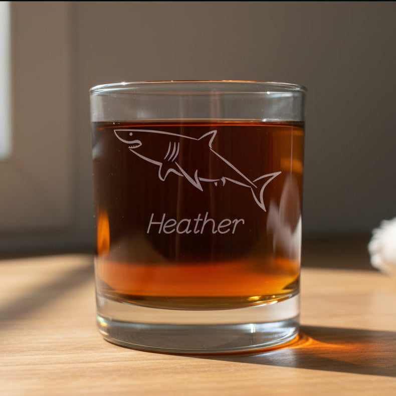 A clear glass rocks tumbler with an etched shark design and the name 'Heather' on the front, filled with amber liquid, set on a wooden surface