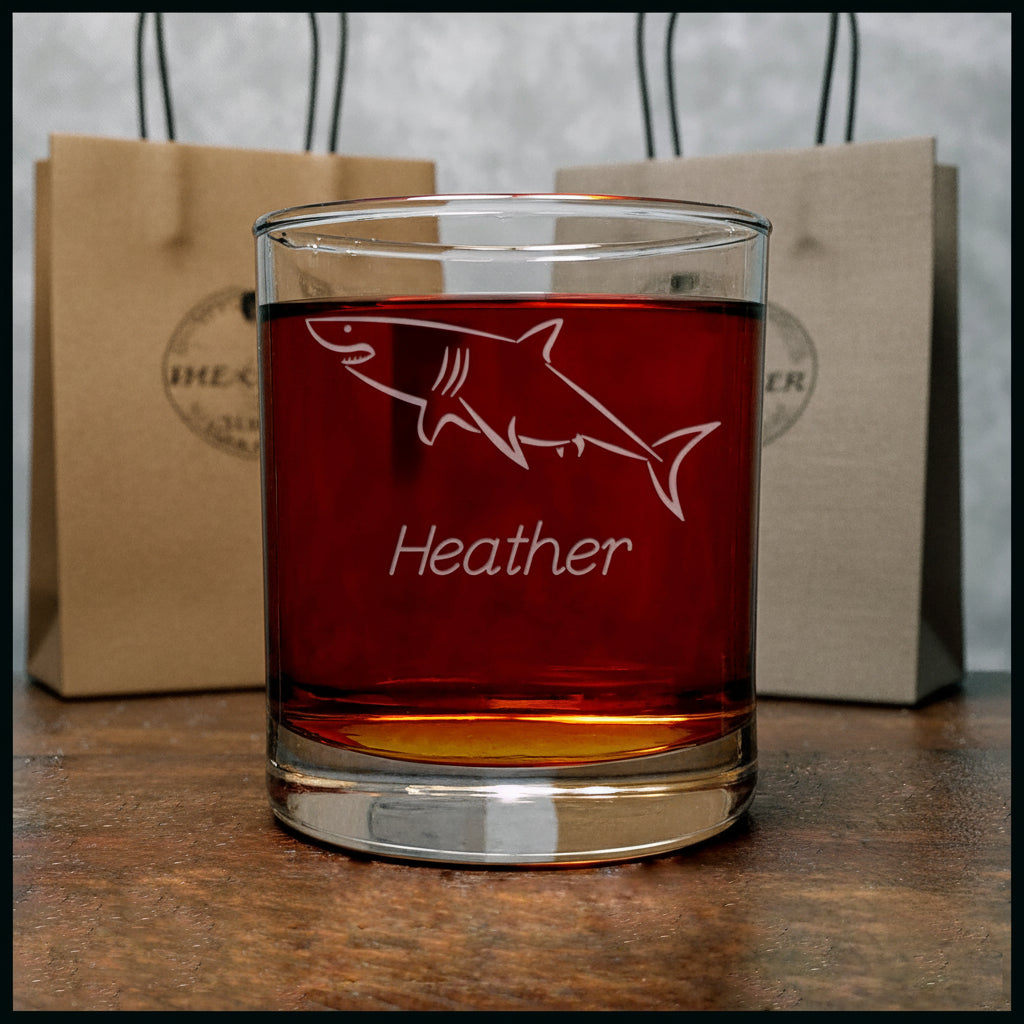 A clear glass rocks tumbler with a white etched shark outline and the name 'Heather' on the front, filled with a dark amber drink and set on a wooden surface against a gray background.