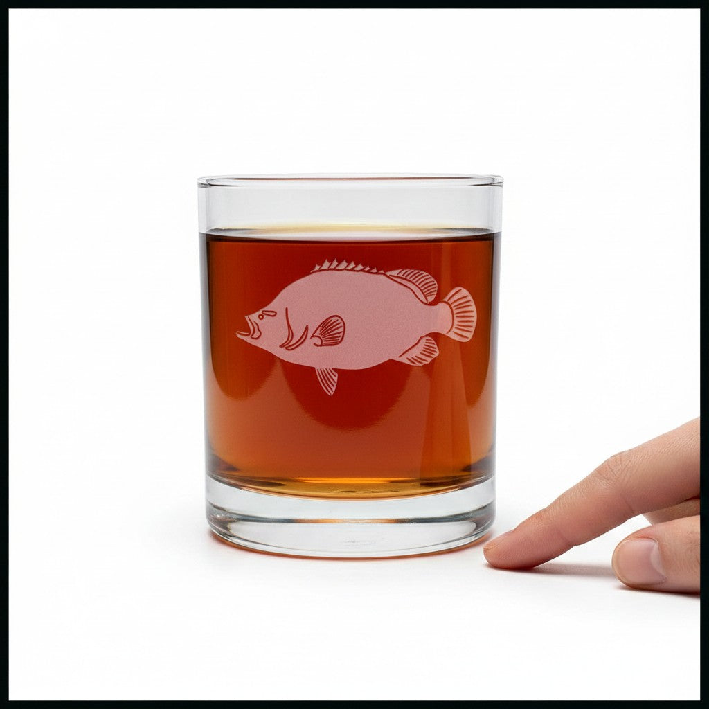 A clear rocks glass filled with amber liquid, featuring a pink fish graphic on the front, with a person's finger pointing at it against a white background.