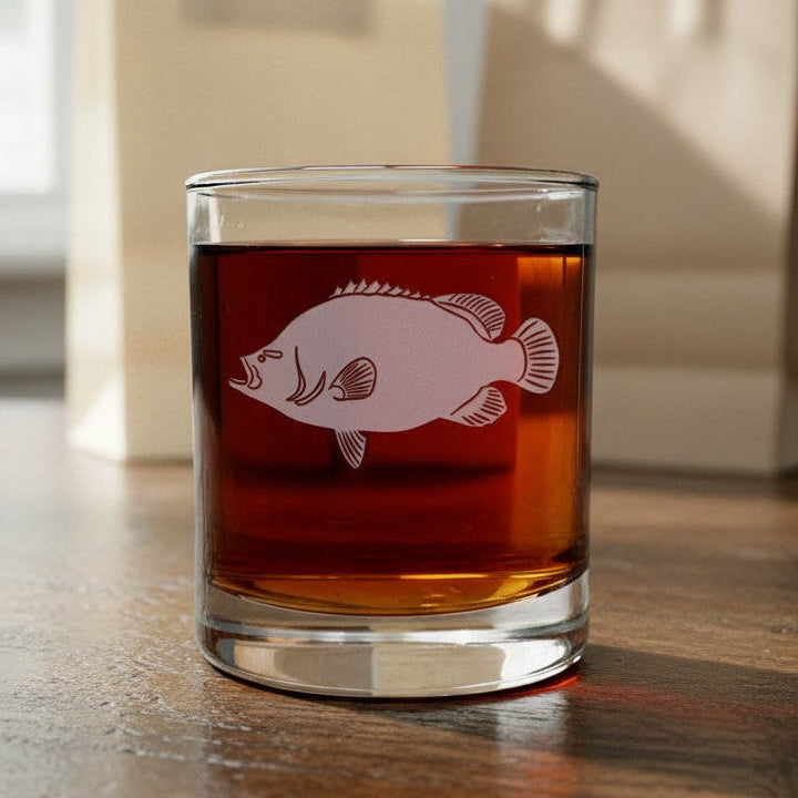 A clear glass tumbler with a white fish illustration sits on a wooden table, filled with amber liquid. Two beige paper shopping bags are blurred in the background near a window.
