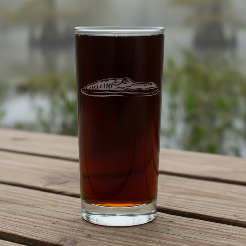 A tall, slender clear glass tumbler filled with a dark liquid, featuring an etched alligator head design on the front, set against a swamp background.