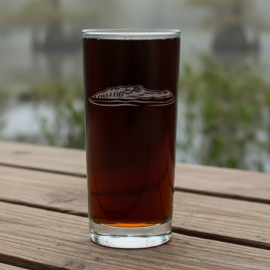 A tall, slender clear glass tumbler filled with a dark liquid, featuring an etched alligator head design on the front, set against a swamp background.