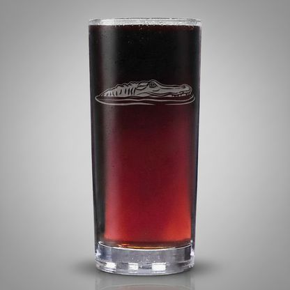 A tall, slender clear glass tumbler filled with a dark red liquid, featuring an  etched alligator design on the front, isolated on a light grey background.