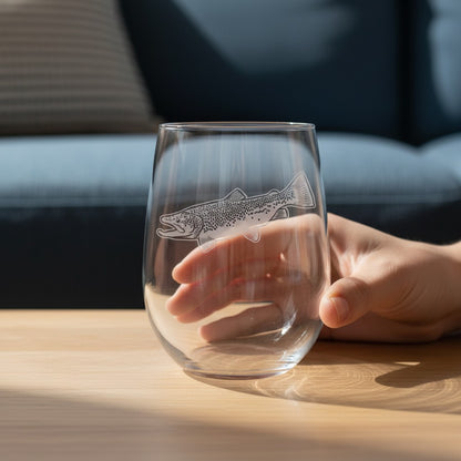 A clear stemless wine glass with an etched trout design, held by a hand on a light wood table in a softly blurred living room setting.