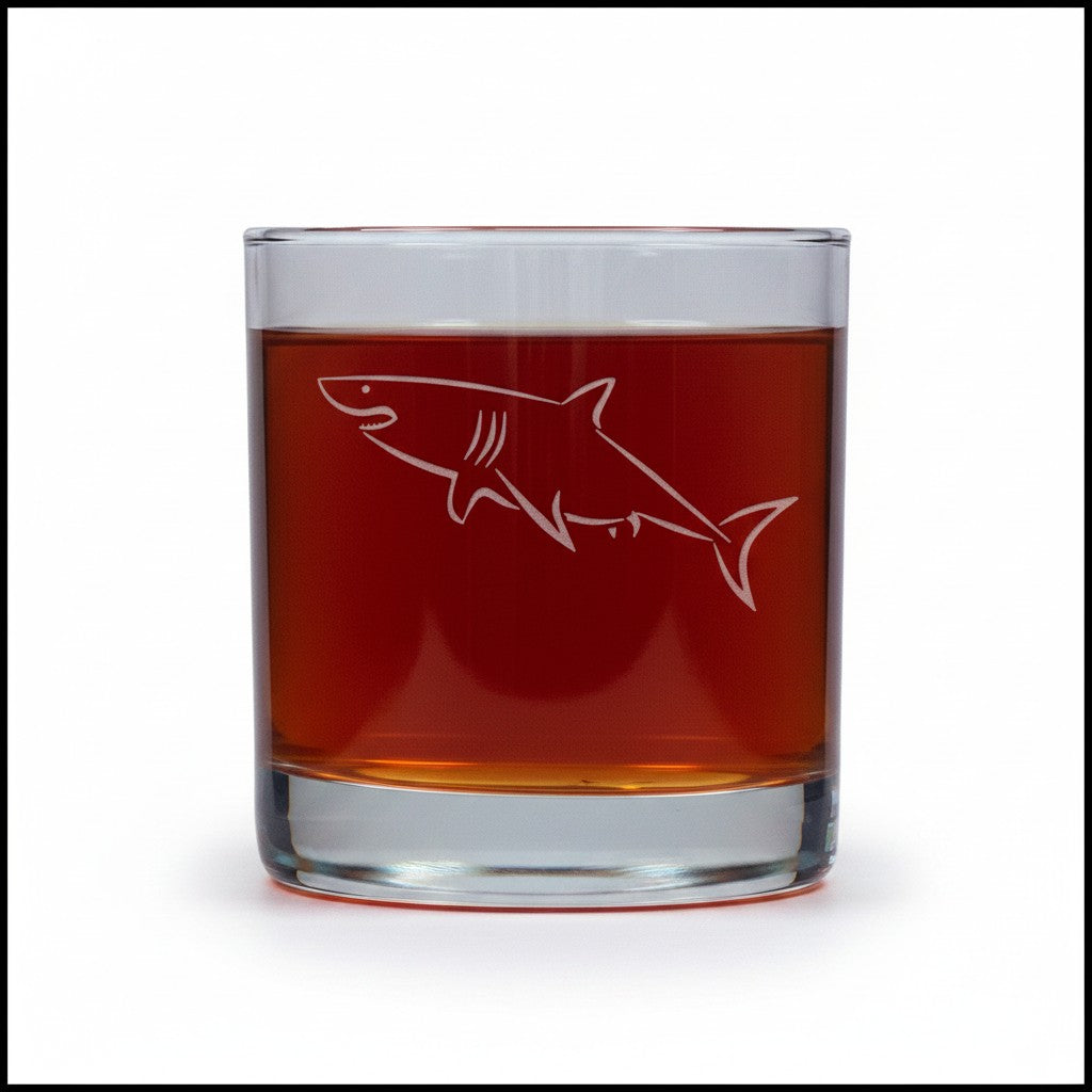 A clear rocks glass containing an amber-brown liquid, with a white etched shark design on the front, shown against a plain white background.