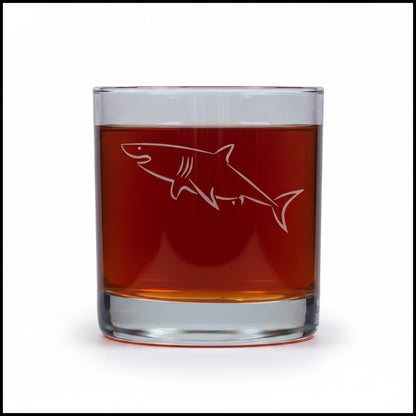 A clear rocks glass containing an amber-brown liquid, with a white etched shark design on the front, shown against a plain white background.