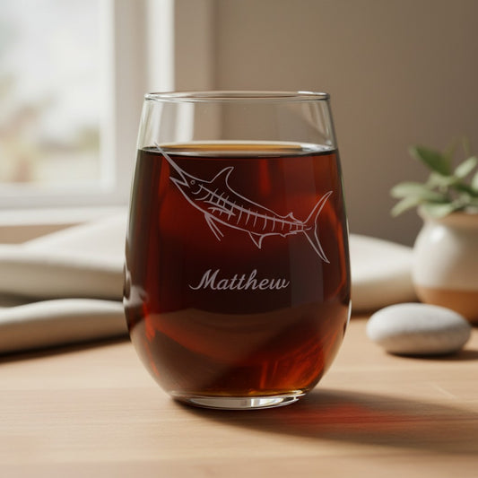 A clear stemless wine glass filled with dark amber liquid, etched with a fish design and the name Matthew, sitting on a wooden table in a softly lit indoor setting.