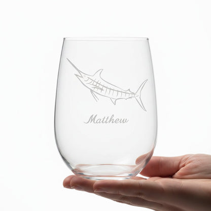 A clear stemless wine glass with an etched sailfish design and the name 'Matthew' below it, held in a person's hand against a white background.