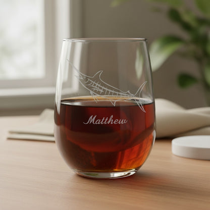 A clear stemless wine glass with an etched marlin and the name "Matthew" on the front, filled with an amber drink and placed on a wooden table in a softly lit indoor setting.