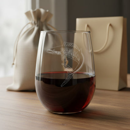 A stemless wine glass filled with red wine, featuring an etched ono fish design on the front, set on a wooden table with a beige drawstring pouch and gift bag in the background.